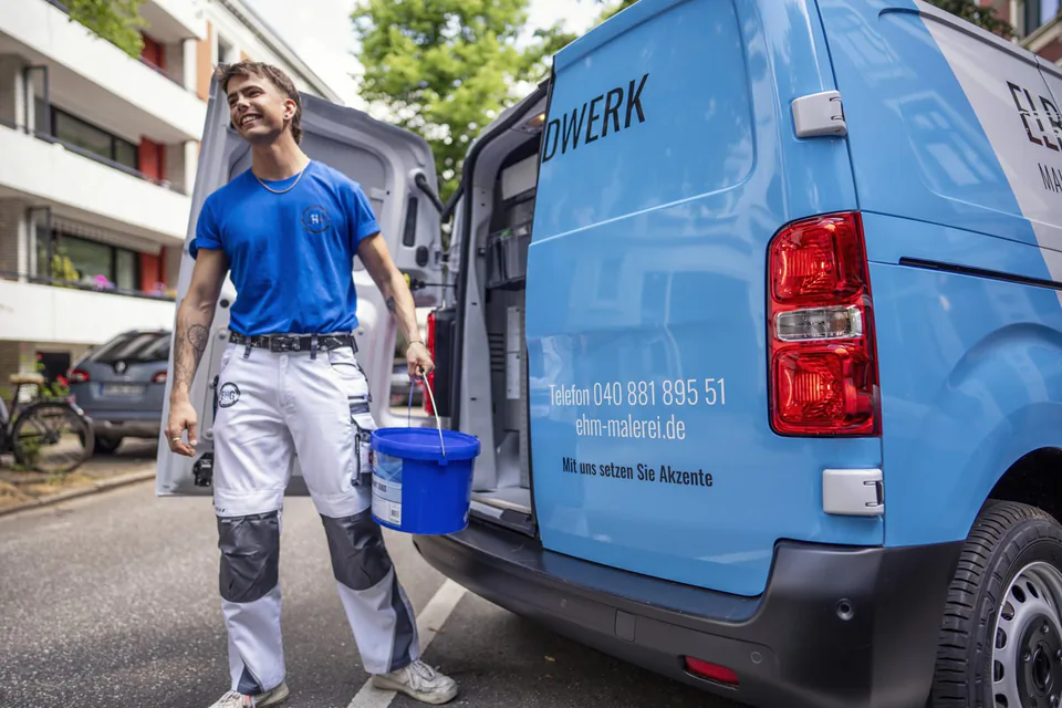 Handwerker mit blauem Eimer neben offenem Firmenwagen von Elb Handwerk auf einer sonnigen Wohnstraße.