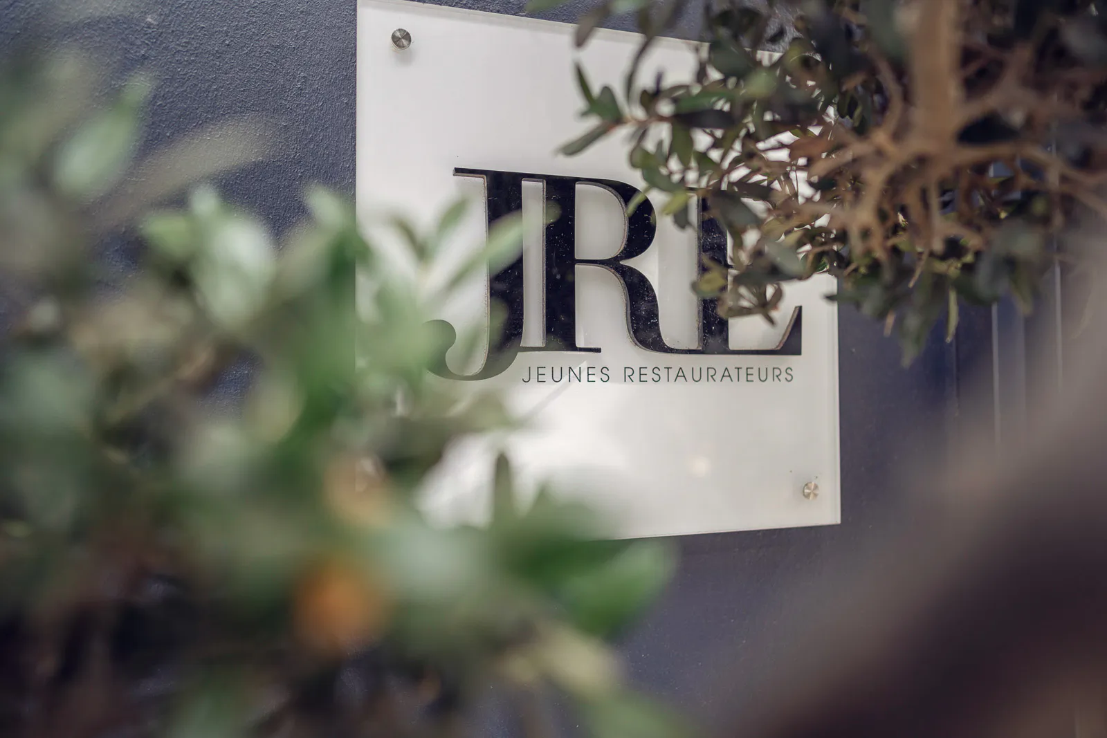 JRE-Schild mit Schriftzug Jeunes Restaurateurs an grauer Außenwand, teilweise von grünen Zweigen verdeckt.