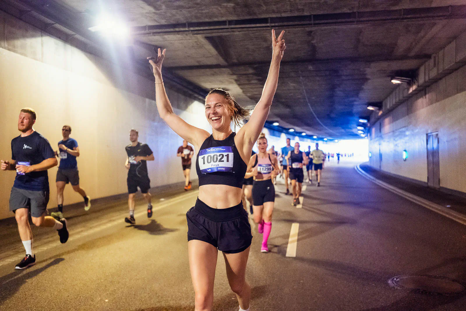 Lächelnde Läuferin mit erhobenen Armen und Startnummer beim Hella Halbmarathon im beleuchteten Tunnel, weitere Läufer.