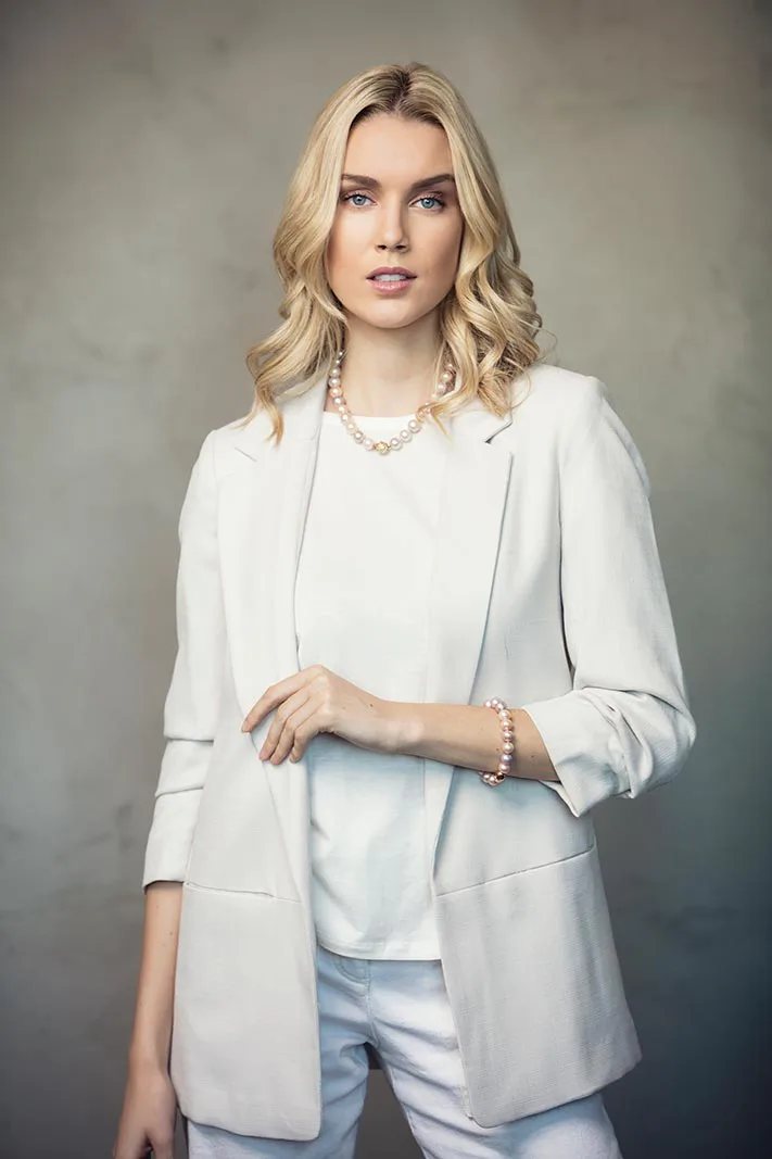 Blonde Frau in weißem Blazer mit Perlenkette und Perlenarmband vor grauer Wand in weichem Licht.