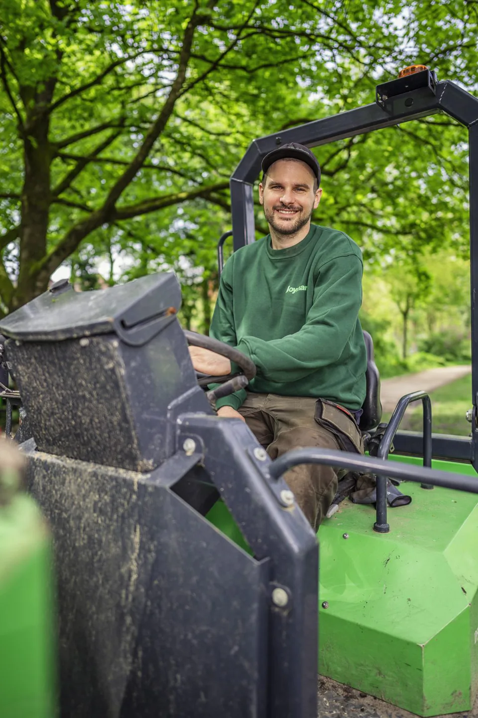 Lächelnder Boymann-Mitarbeiter in grünem Pullover sitzt auf einer grünen Arbeitsmaschine unter Bäumen.