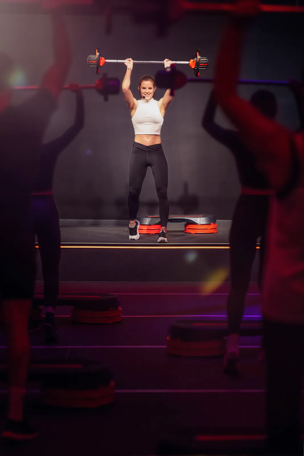 Frau hebt eine Langhantel über den Kopf, vor ihr trainiert eine Gruppe im rot-violett beleuchteten Fitnessraum.