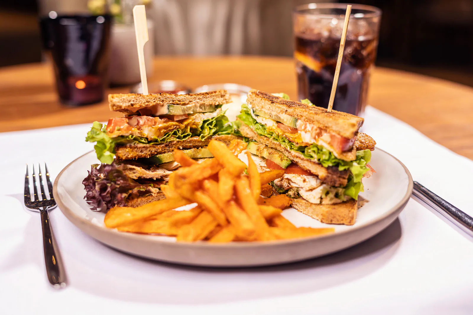 Club-Sandwich mit Salat, Speck und Pommes auf einem Teller im Restaurant, dahinter ein dunkles Getränk im warmen Licht.