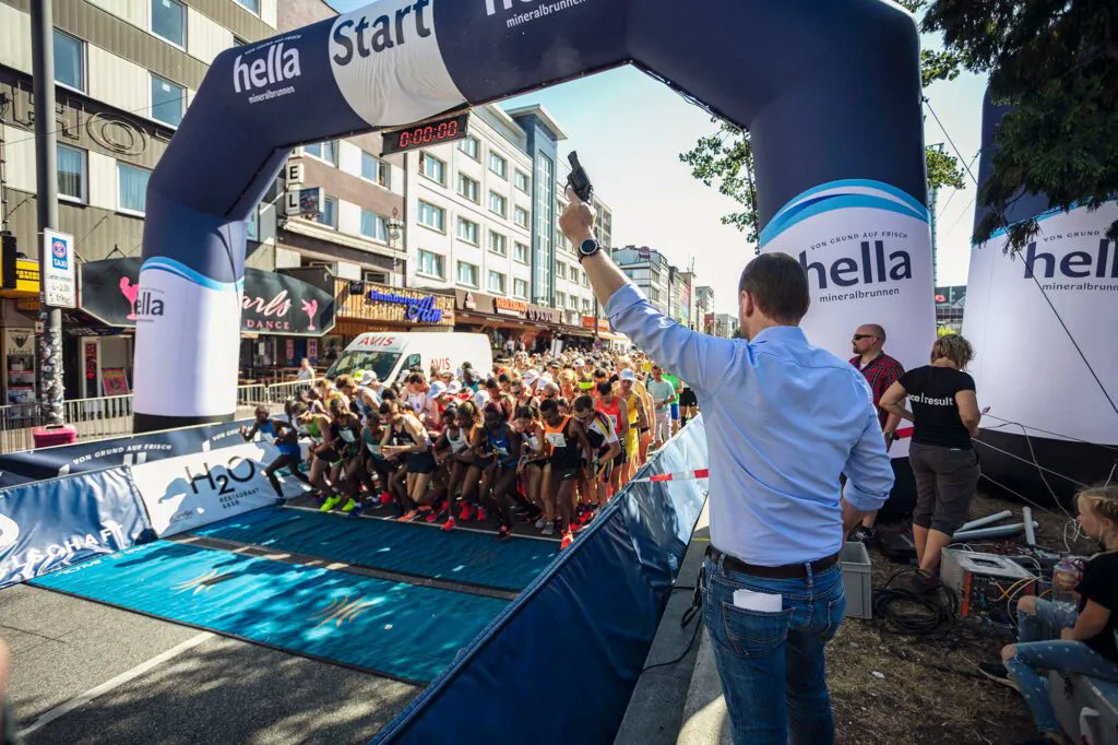 Ein Mann hebt die Startpistole, während Läufer unter dem hella-Startbogen auf einer sonnigen Straße loslaufen.