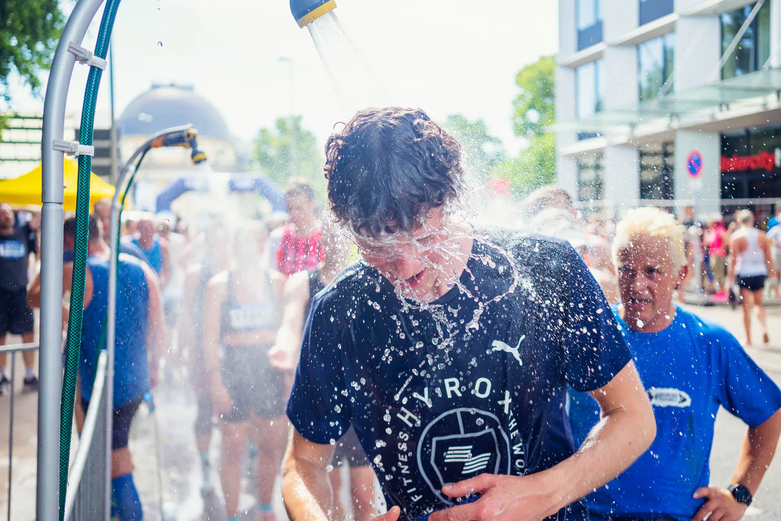 Läufer kühlt sich beim Hella Halbmarathon unter einer Wasserdusche auf sonniger Straße ab, weitere Sportler dahinter.