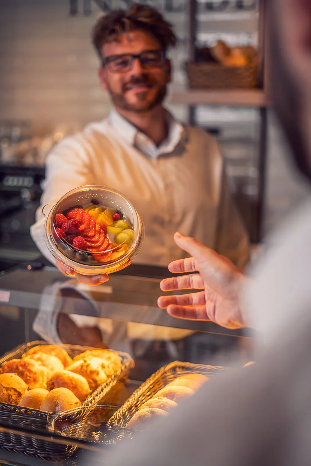 Mann mit Brille reicht eine Schale mit frischem Obst über eine Glastheke mit Brötchenkörben im warmen Licht.