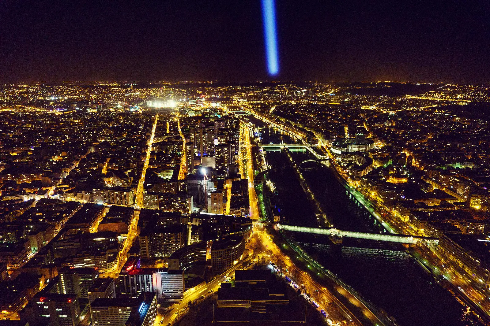 Nächtlicher Blick über Paris mit beleuchteten Straßen, Brücken über die Seine und blauem Lichtreflex am Himmel.