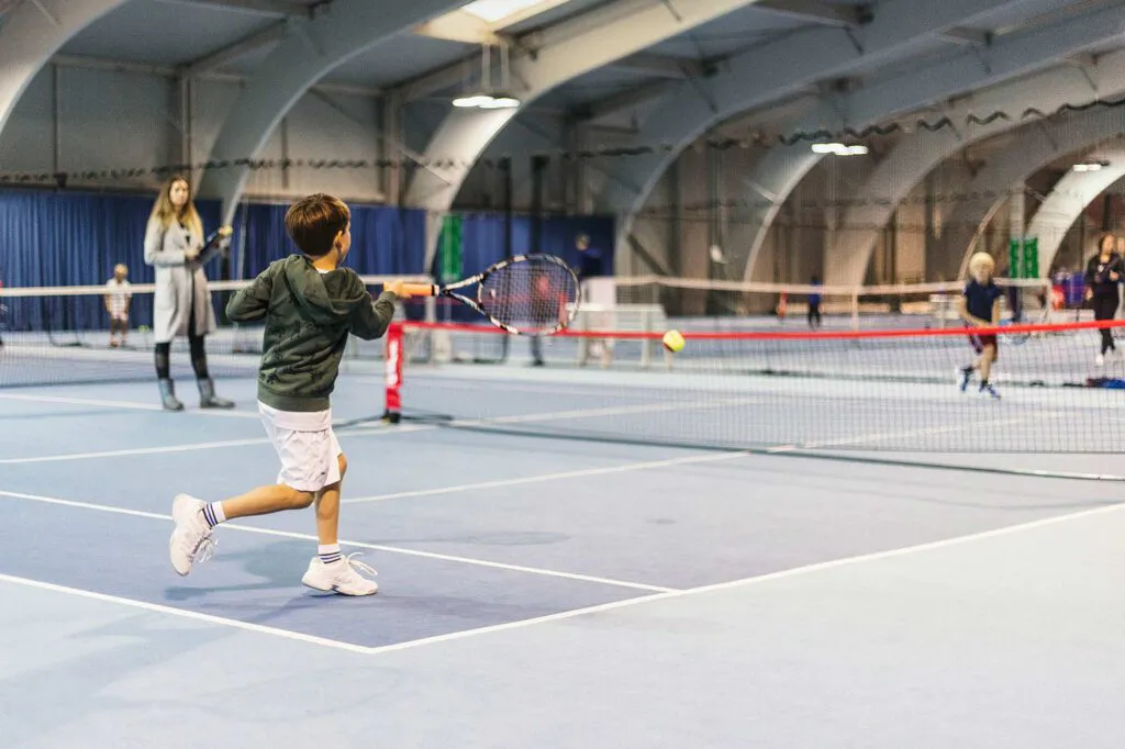 Kind schlägt einen Tennisball über ein niedriges Netz in einer beleuchteten Tennishalle beim Wilson Master Cup.