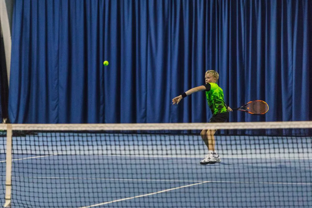 Junger Tennisspieler im grünen Shirt holt in einer Halle mit blauem Vorhang zur Vorhand aus, der Ball fliegt übers Netz.