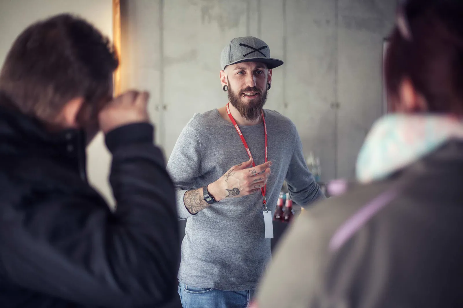 Bärtiger Mann mit grauer Kappe und rotem Umhängeband gestikuliert im Gespräch mit zwei Personen vor einer Betonwand.