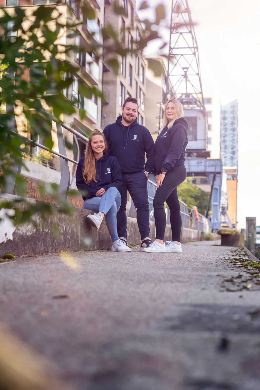 Drei Waterkant-Teammitglieder in dunklen Hoodies posieren im Sonnenlicht vor einem Backsteingebäude und einem Kran.