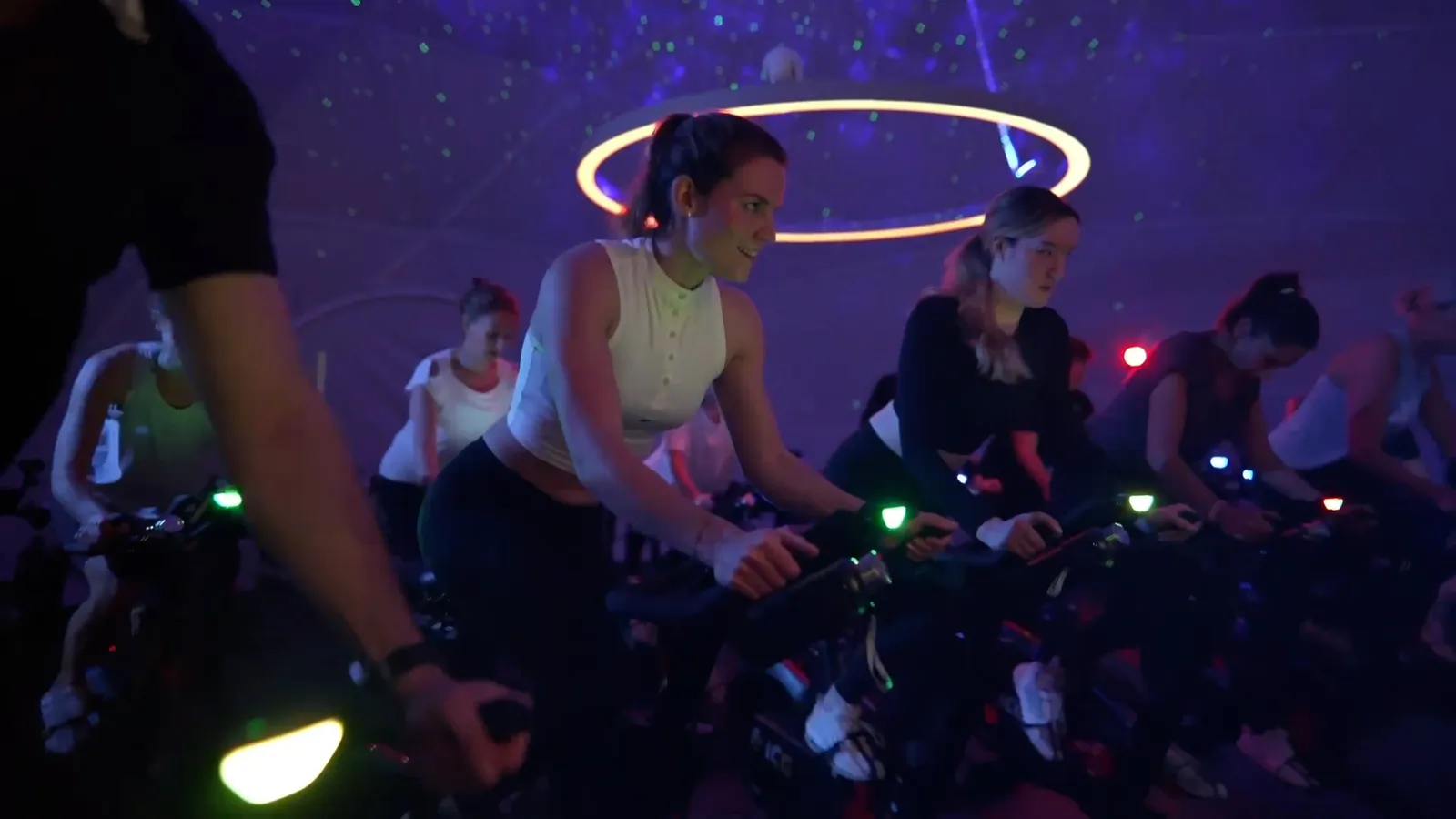 Gruppe trainiert auf Indoor-Bikes in einem dunklen Raum mit violettem Licht und leuchtendem Deckenring.