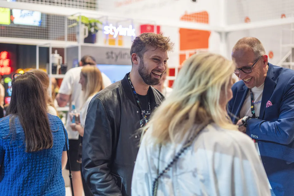 Lachende Messebesucher im Gespräch am hell beleuchteten Stand, eine blonde Frau im Vordergrund.