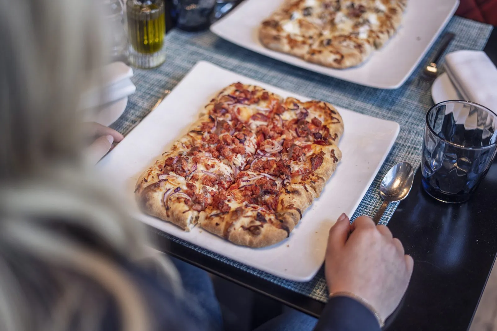 Person mit Besteck vor einer länglichen Pizza mit roten Zwiebeln auf weißem Teller am gedeckten Tisch.