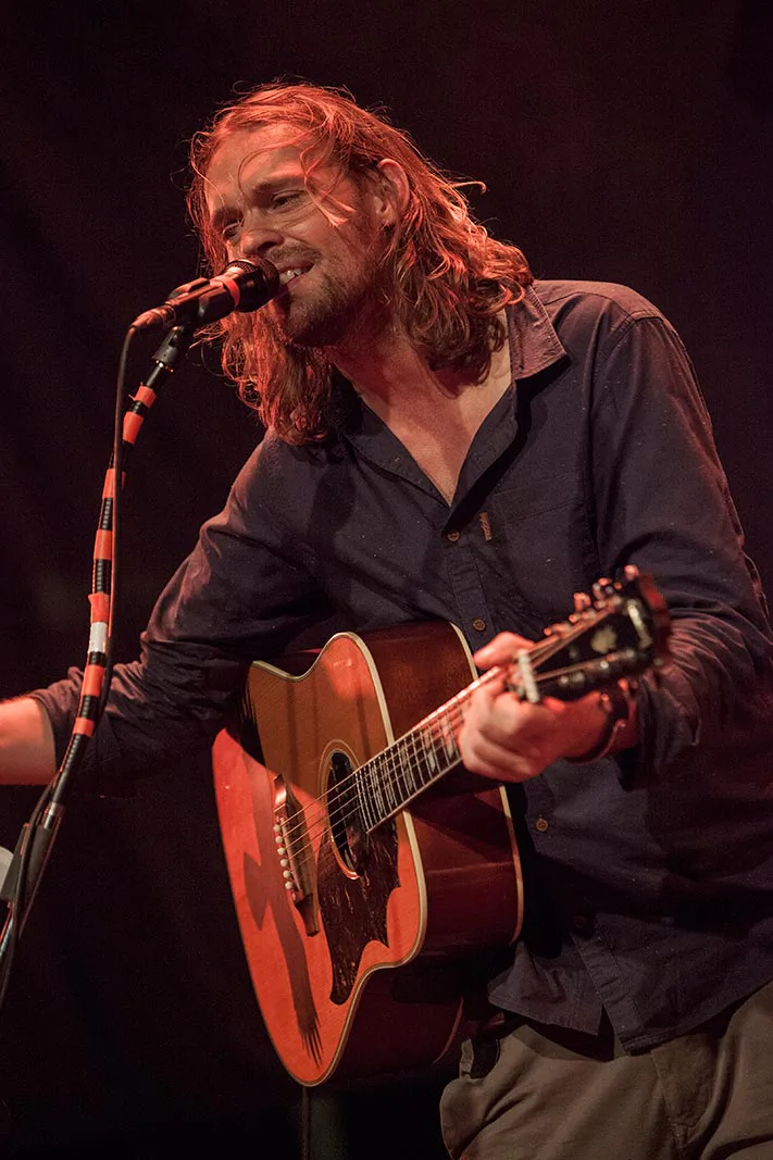 Ingo Pohlmann singt am Mikrofon und spielt Akustikgitarre auf einer Bühne im roten Licht.