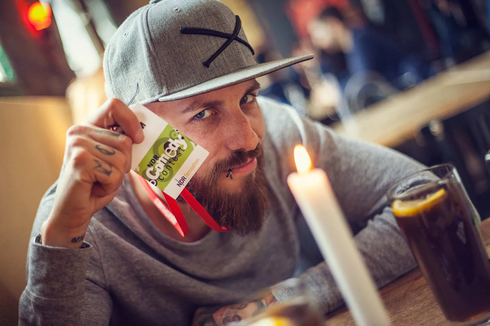 Dennis Grundt mit grauer Kappe hält einen NDR-Comedy-Contest-Ausweis am Tisch; davor eine Kerze und ein Getränk.
