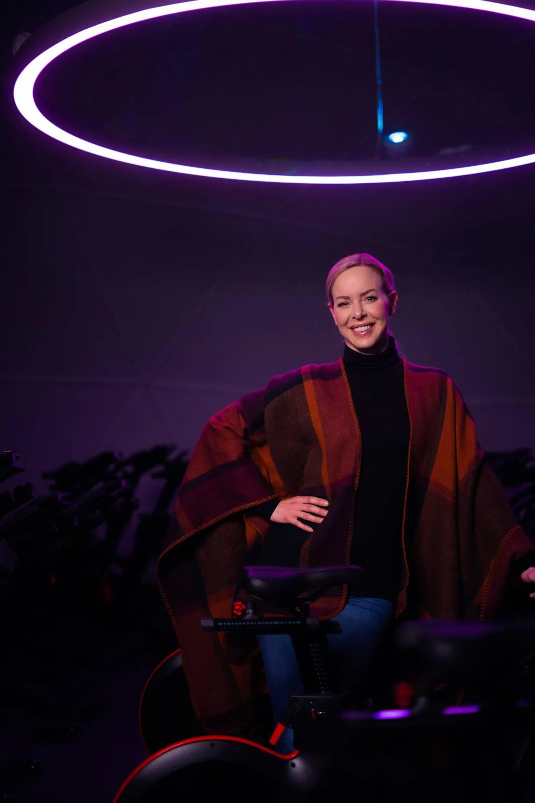 Isabel Edvardsson posiert lächelnd zwischen Indoor-Bikes unter einem violett leuchtenden Deckenring.