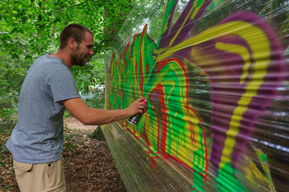 Mann sprüht buntes Graffiti auf eine zwischen Bäumen gespannte transparente Folie.