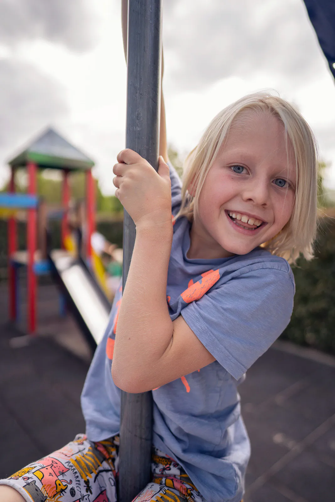 Lächelndes blondes Kind hält sich bei Tageslicht an einer Stange auf einem Spielplatz mit Rutsche fest.