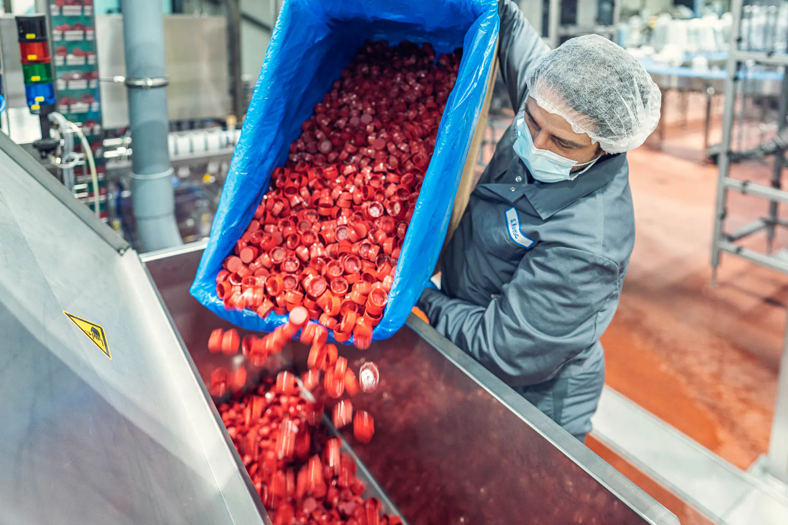 Mitarbeiter mit Haarnetz und Mundschutz schüttet rote Flaschenverschlüsse in einen Metalltrichter der Abfüllanlage.
