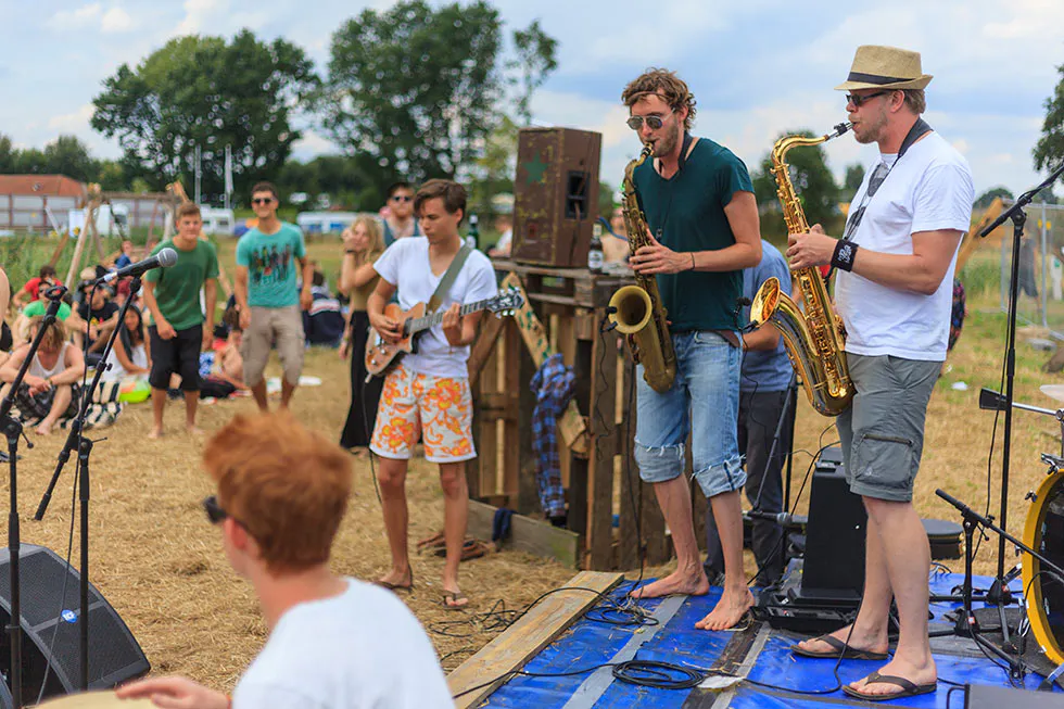 Zwei Saxofonisten und ein Gitarrist spielen barfuß auf einer kleinen Freiluftbühne vor Publikum.