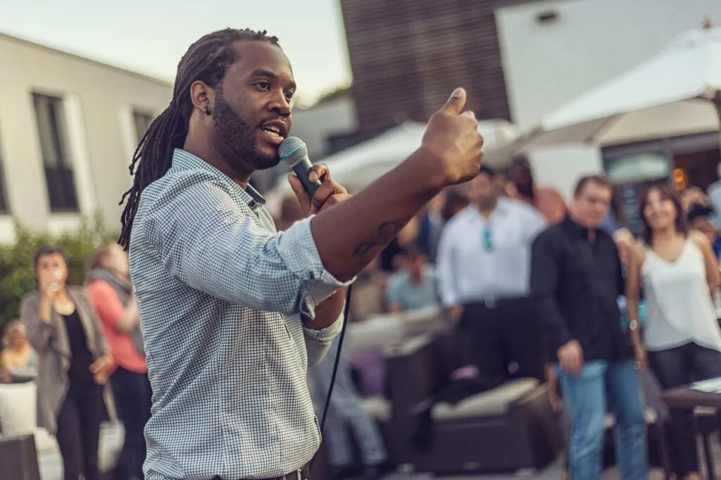 Sänger mit Dreadlocks hält ein Mikrofon und zeigt den Daumen hoch vor Publikum auf einer Terrasse bei Tageslicht.