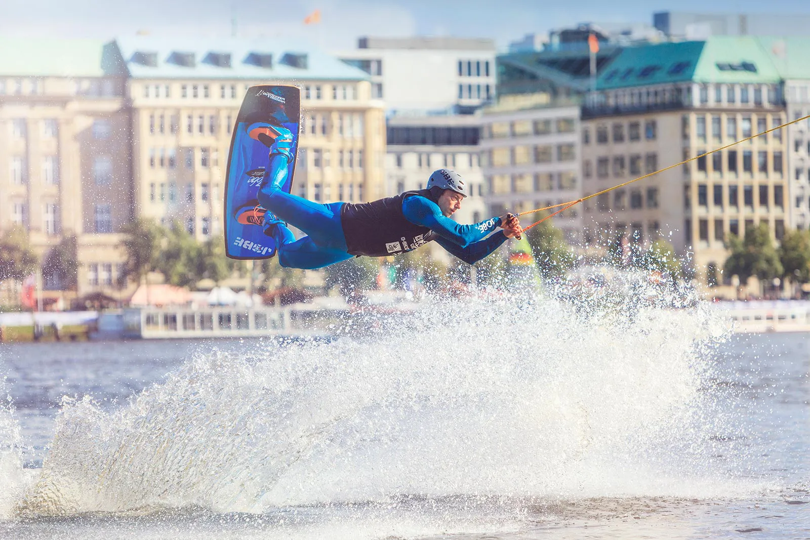 Wakeboarder mit Kappe springt am Zugseil über das Wasser, vor einer sonnigen Stadtkulisse mit hoher Gischt.