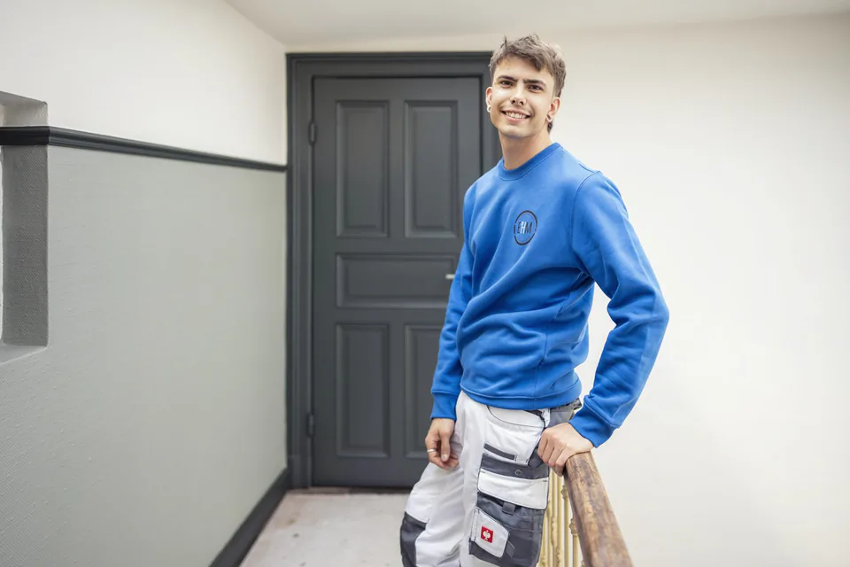 Lächelnder Handwerker im blauen Pullover und weißer Arbeitshose am Treppengeländer vor einer dunklen Tür.