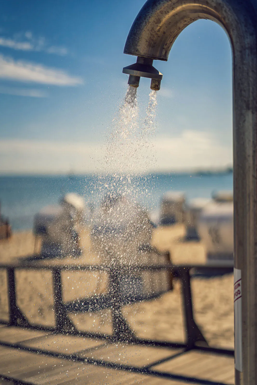 Wasser sprüht aus einer gebogenen Stranddusche im Sonnenlicht, dahinter Sand, Strandkörbe und Meer.