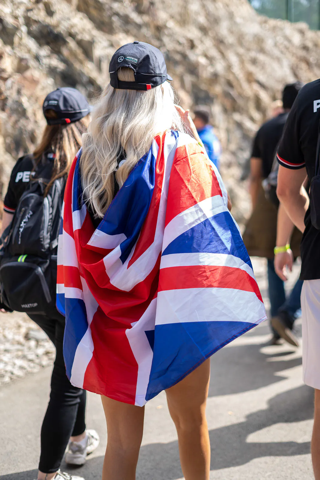 Person mit langen blonden Haaren, schwarzer Kappe und britischer Flagge als Umhang auf sonnigem Weg neben Felswand.