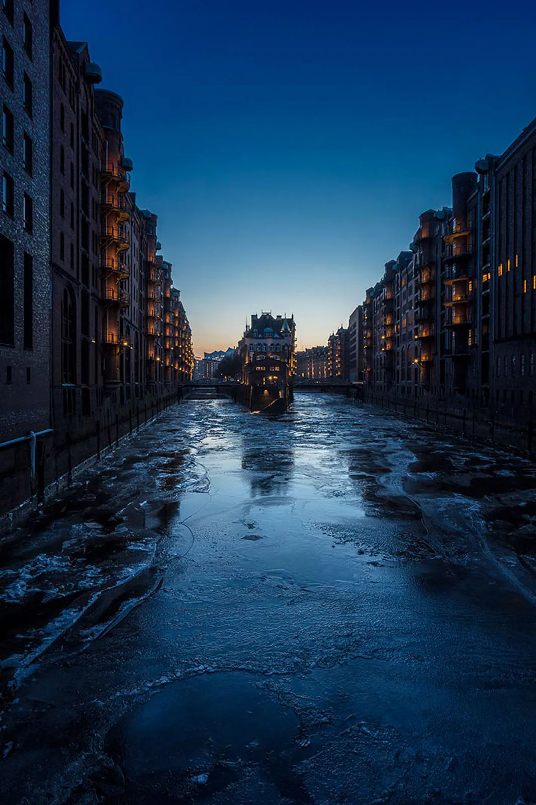 Vereister Kanal zwischen hohen Backsteinspeichern in der blauen Dämmerung, warme Lichter in den Fenstern.