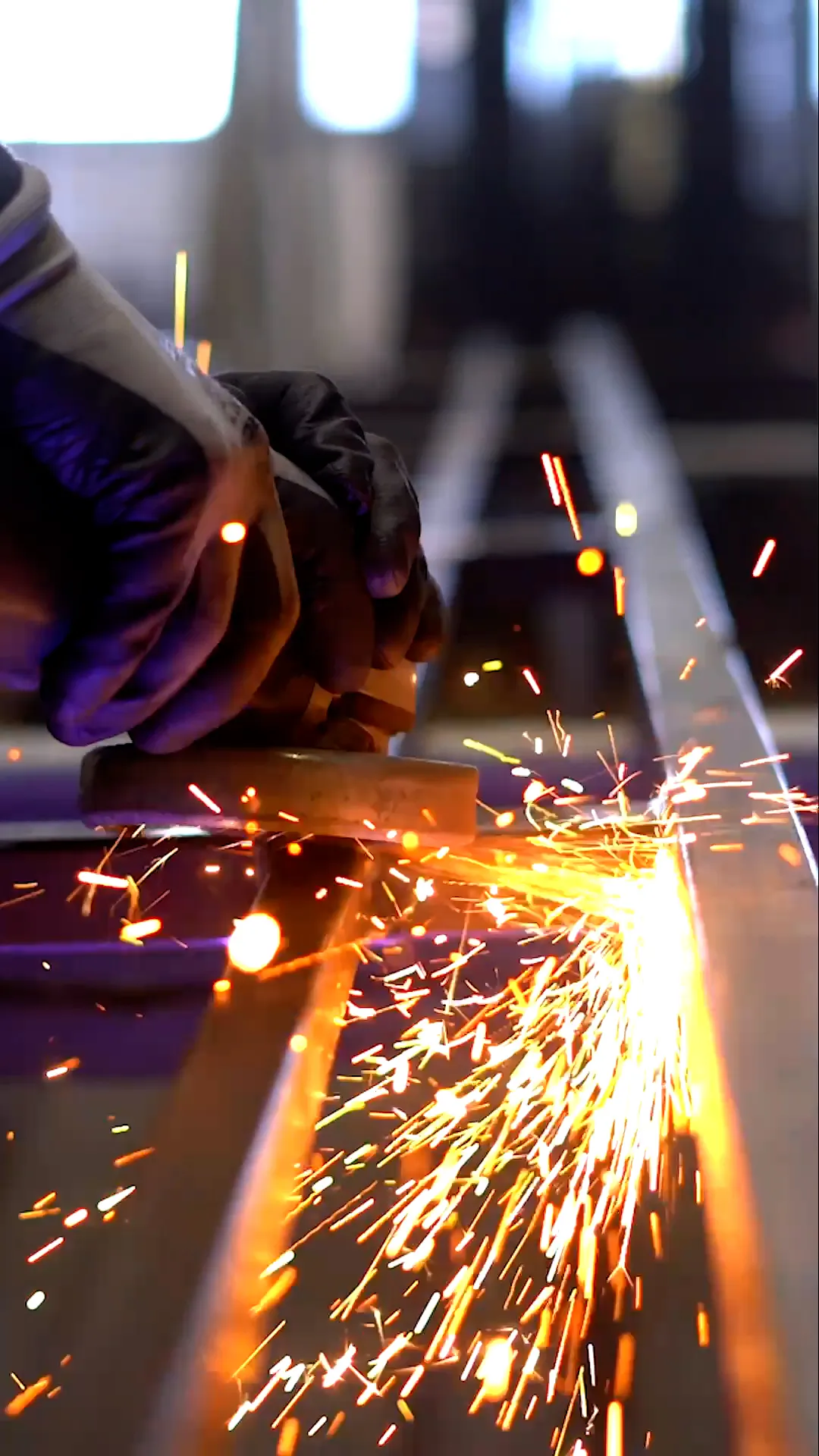 Hände mit Arbeitshandschuhen schleifen ein Metallprofil in einer Werkstatt, helle Funken sprühen.