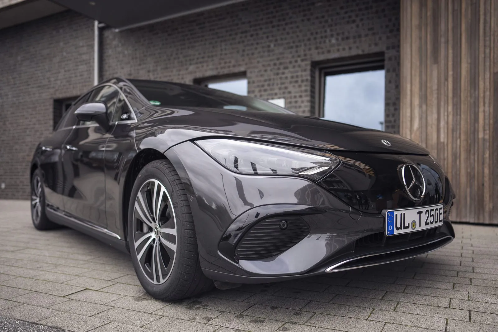 Dunkler Mercedes-Benz in Front- und Seitenansicht auf gepflastertem Hof vor einer Backstein- und Holzfassade.