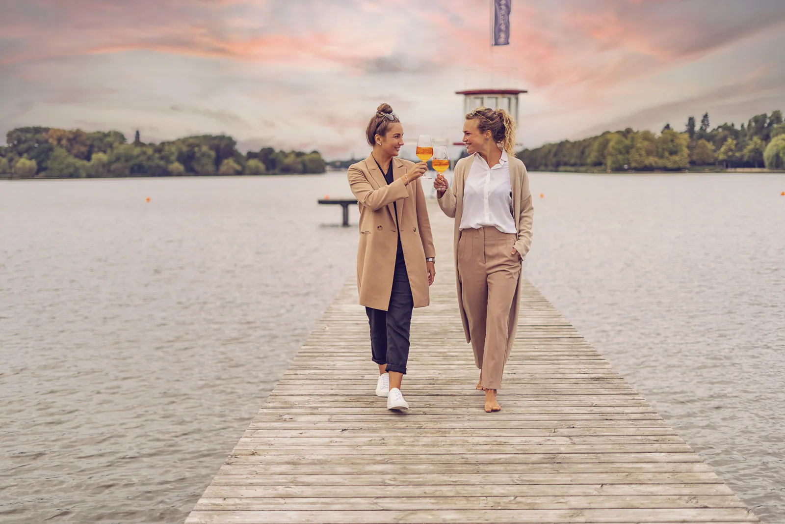 Zwei Frauen stoßen mit orangefarbenen Drinks auf einem Holzsteg am See unter einem rosa Abendhimmel an.