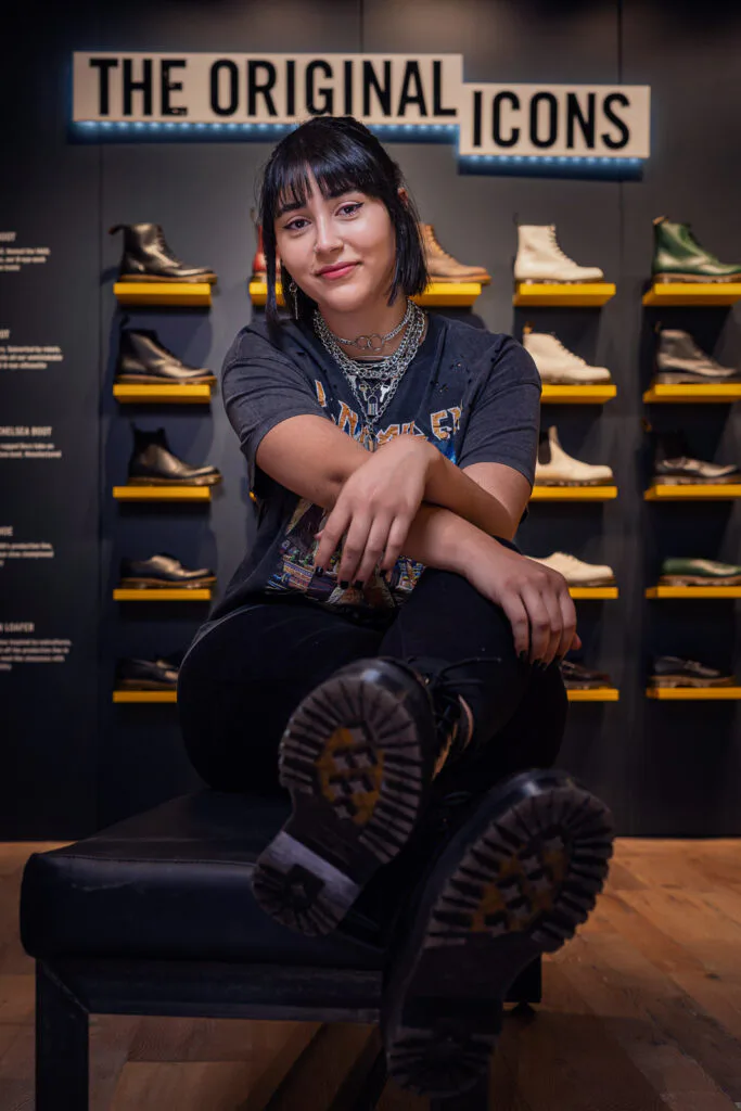 Mitarbeiterin sitzt im Dr. Martens-Laden vor beleuchteten Schuhregalen, die Sohlen ihrer schwarzen Stiefel zur Kamera.
