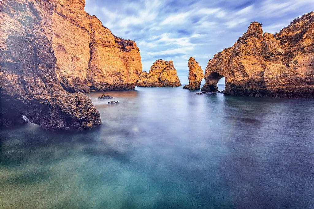 Goldene Felsklippen mit Felsentor an der Algarve, davor glatt belichtetes Meer unter verwischten Wolken.