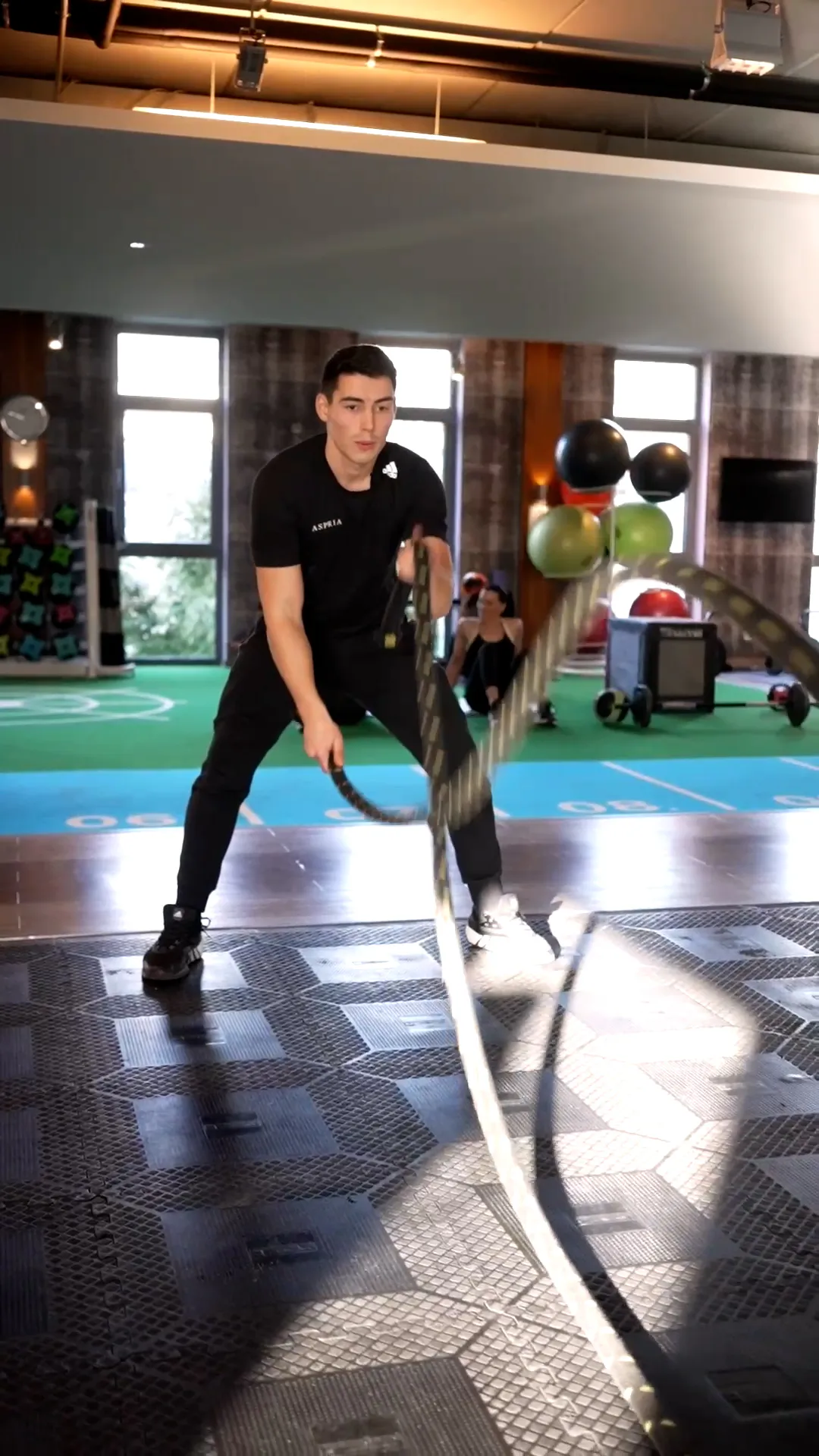 Mann in schwarzer Sportkleidung trainiert mit Battle Ropes im Fitnessstudio, Sonnenlicht fällt durch große Fenster.