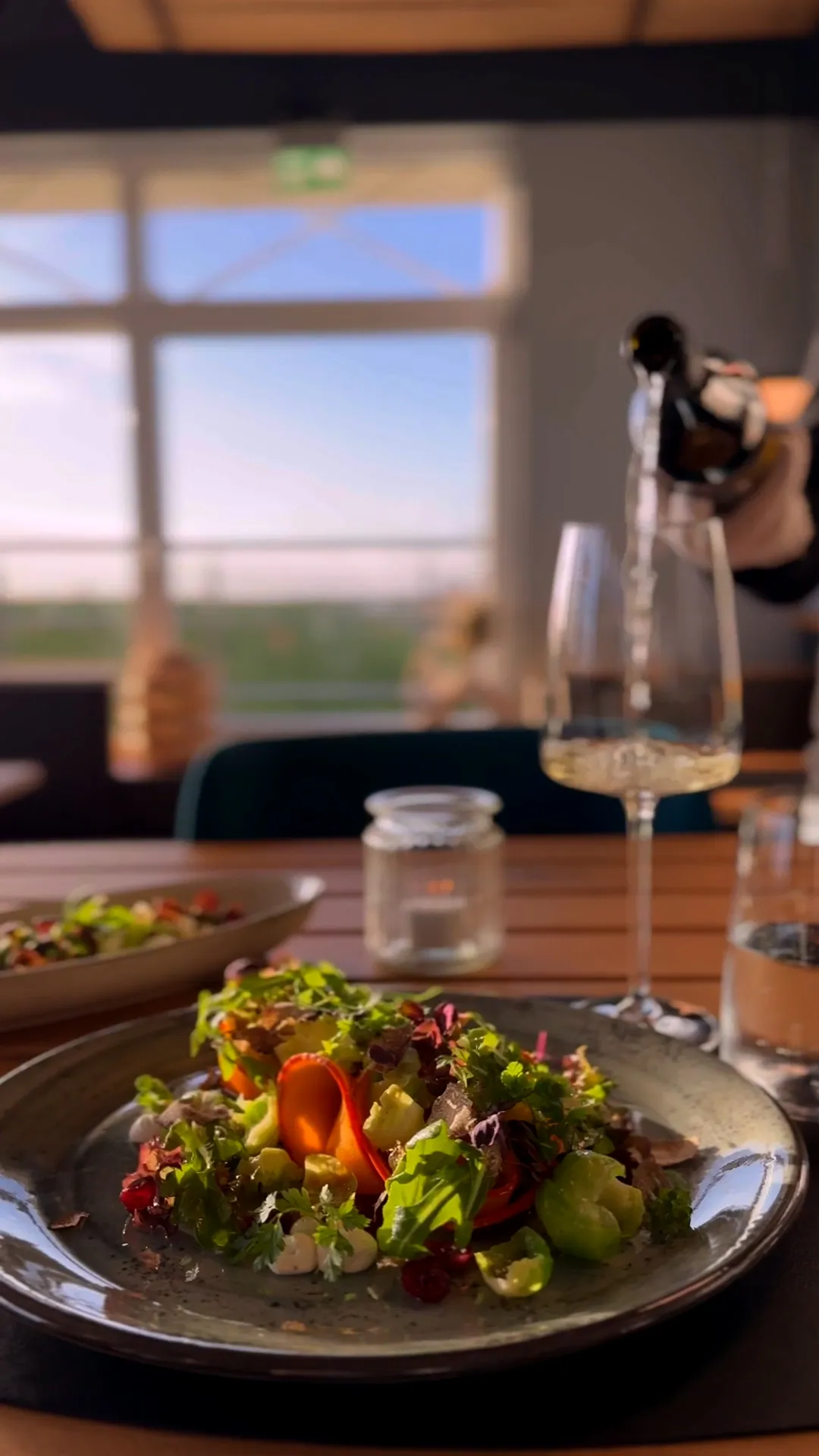 Salat auf einem Keramikteller im warmen Restaurantlicht, dahinter wird Sekt in ein Glas eingeschenkt.