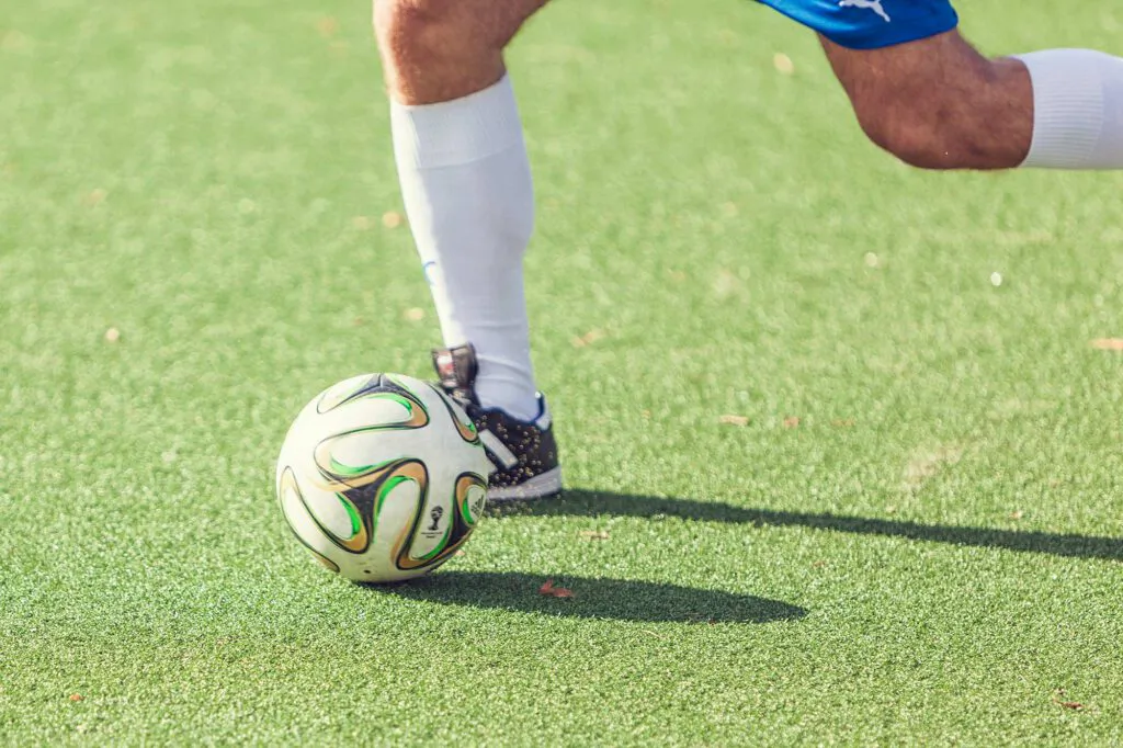 Beine eines Fußballspielers in weißen Stutzen und blauer Hose am Ball auf sonnenbeschienenem Kunstrasen.