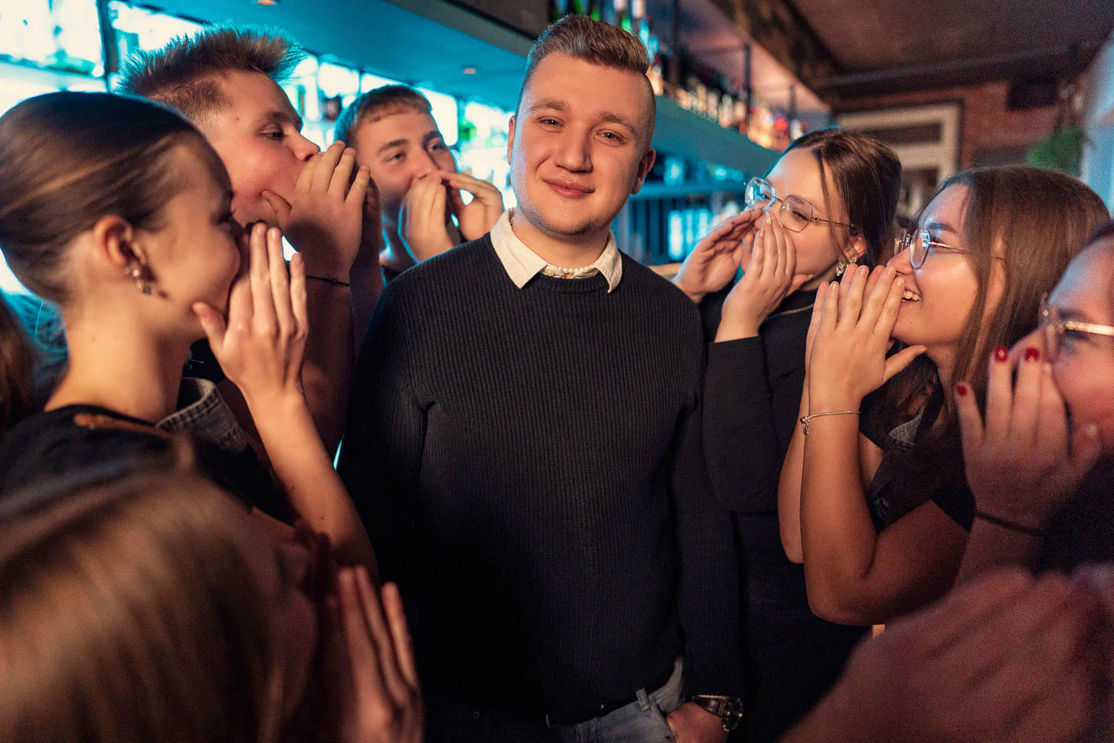 Ein lächelnder Mann steht in einer Restaurantbar, umringt von jungen Leuten mit Händen am Mund, bei türkiswarmem Licht.