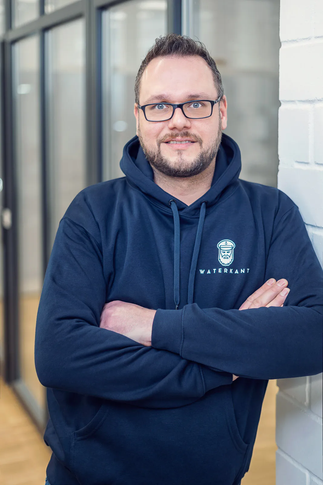 Mann mit Brille im dunkelblauen Waterkant-Hoodie steht mit verschränkten Armen neben einer weißen Backsteinwand.