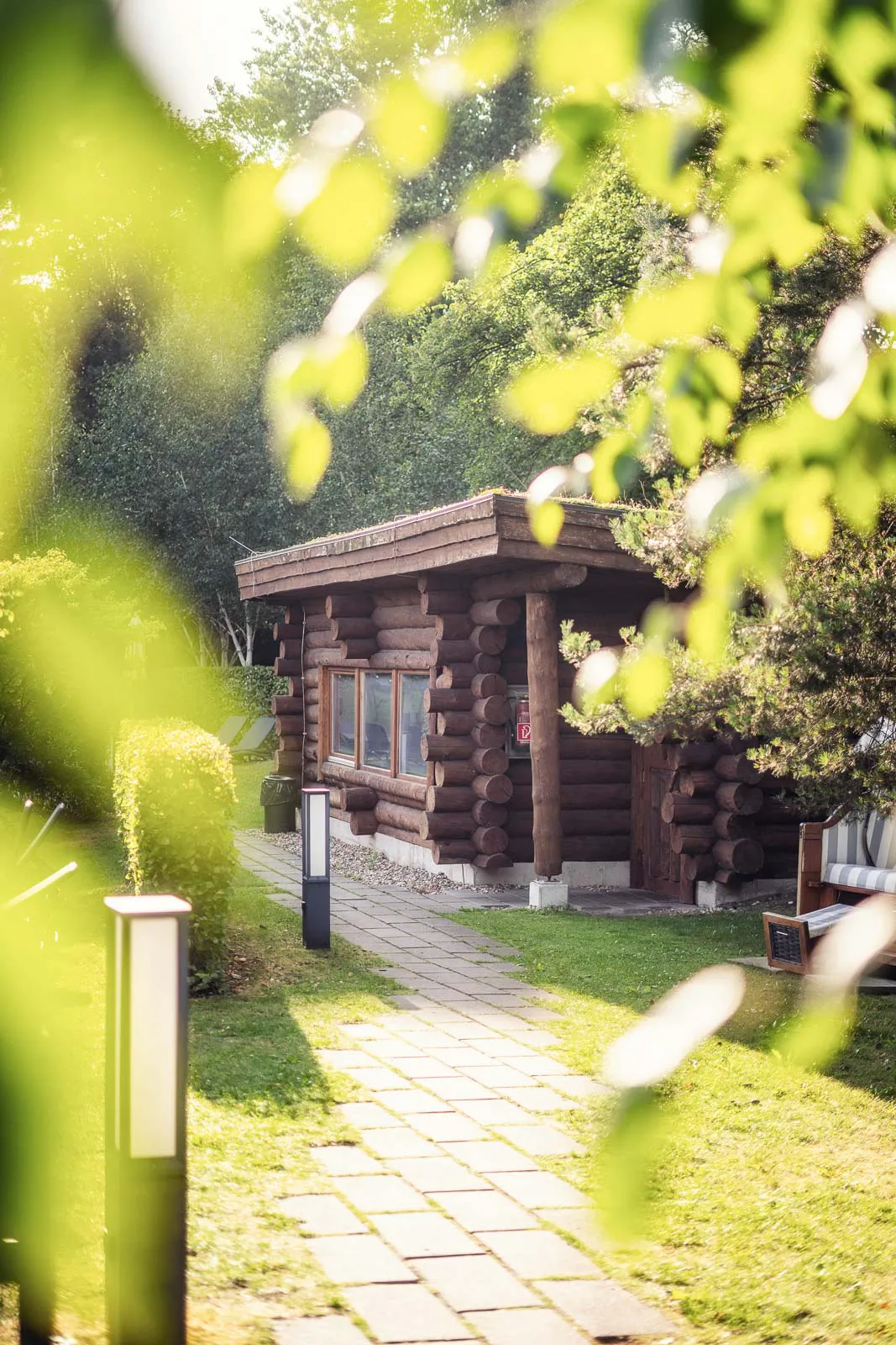 Rustikales Sauna-Blockhaus im sonnigen Garten, davor ein gepflasterter Weg mit Leuchten, umrahmt von grünen Blättern.