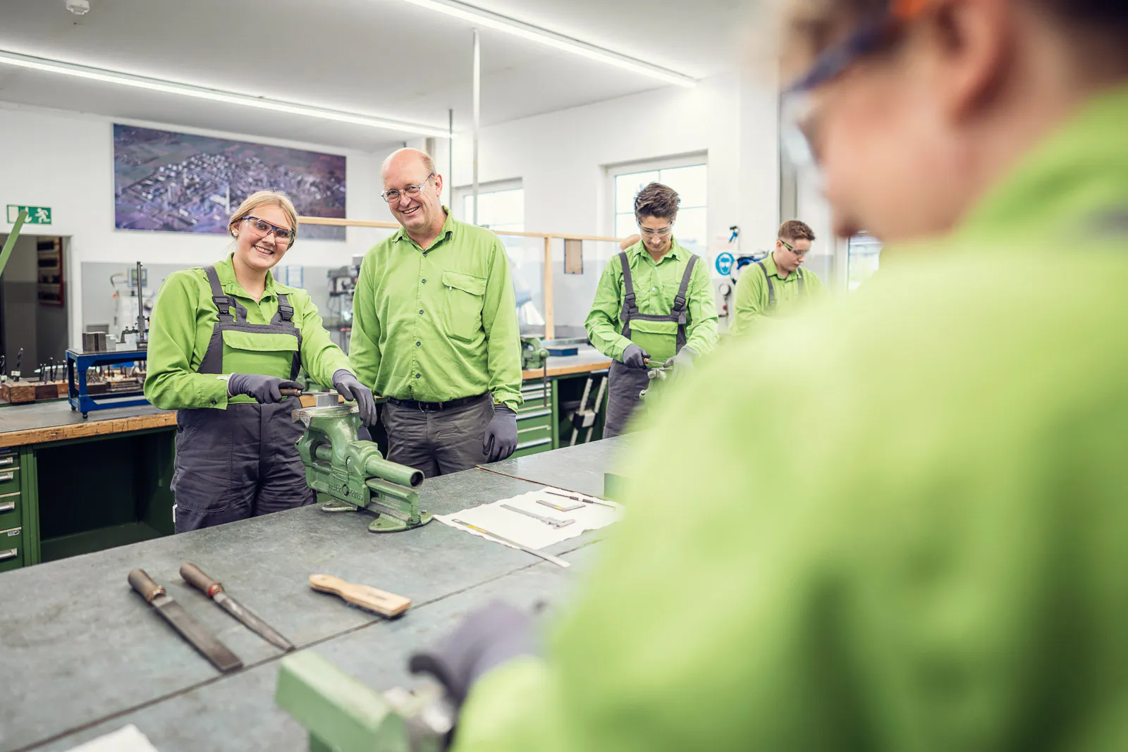 Personen in grüner Arbeitskleidung und Schutzbrillen arbeiten an Schraubstöcken in einer hellen Werkstatt.