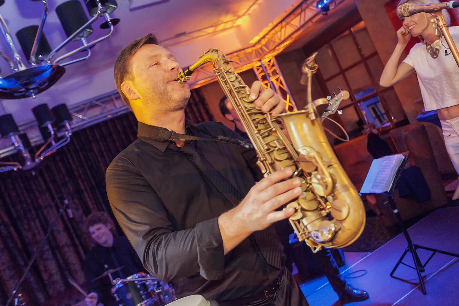 Saxofonist spielt bei warmem Bühnenlicht im Innenraum, dahinter ein Schlagzeuger und eine Frau am Mikrofon.