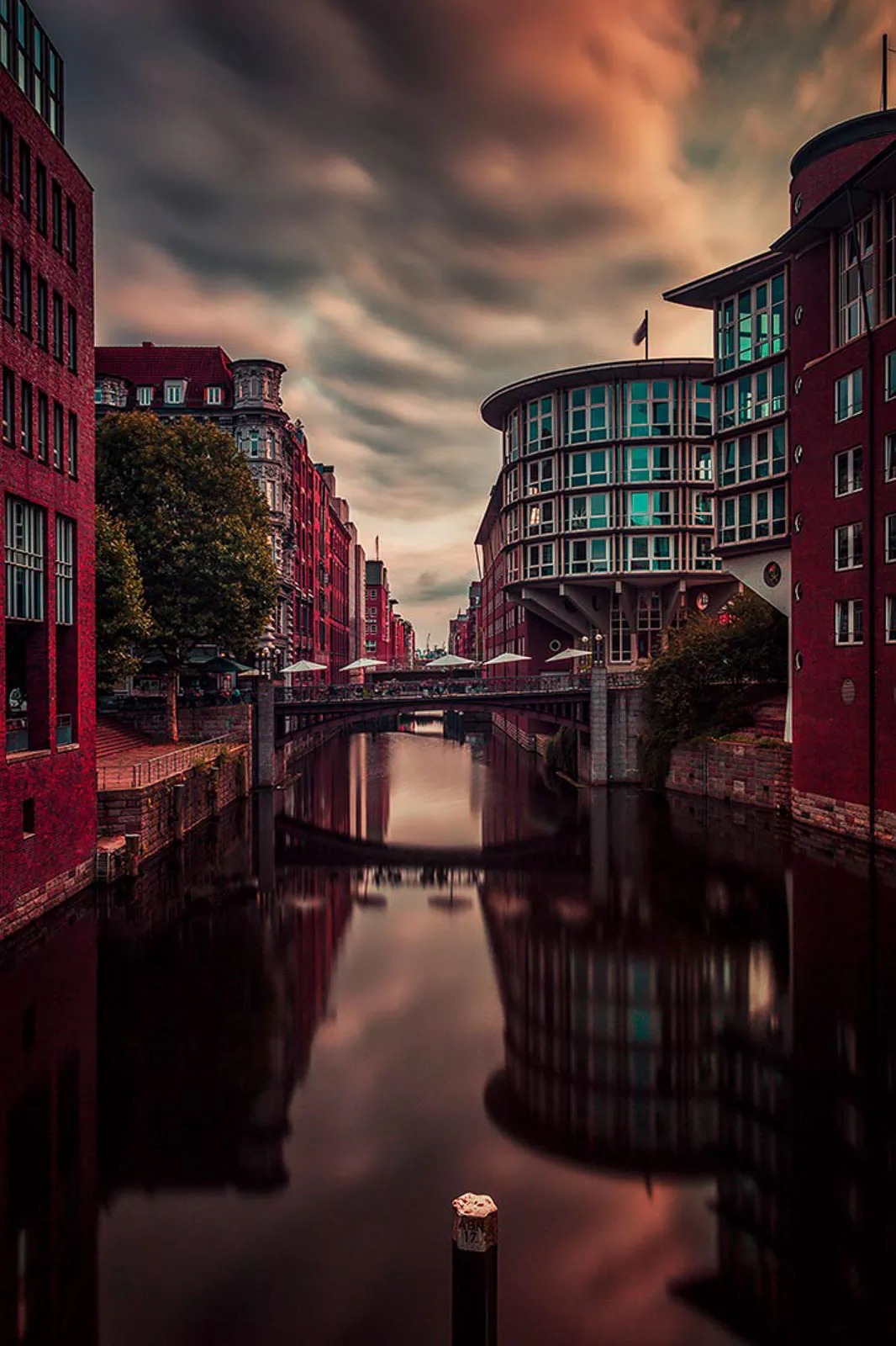 Kanal zwischen roten Backsteingebäuden mit Brücke und Spiegelungen im Wasser unter orangegrauem Wolkenhimmel.