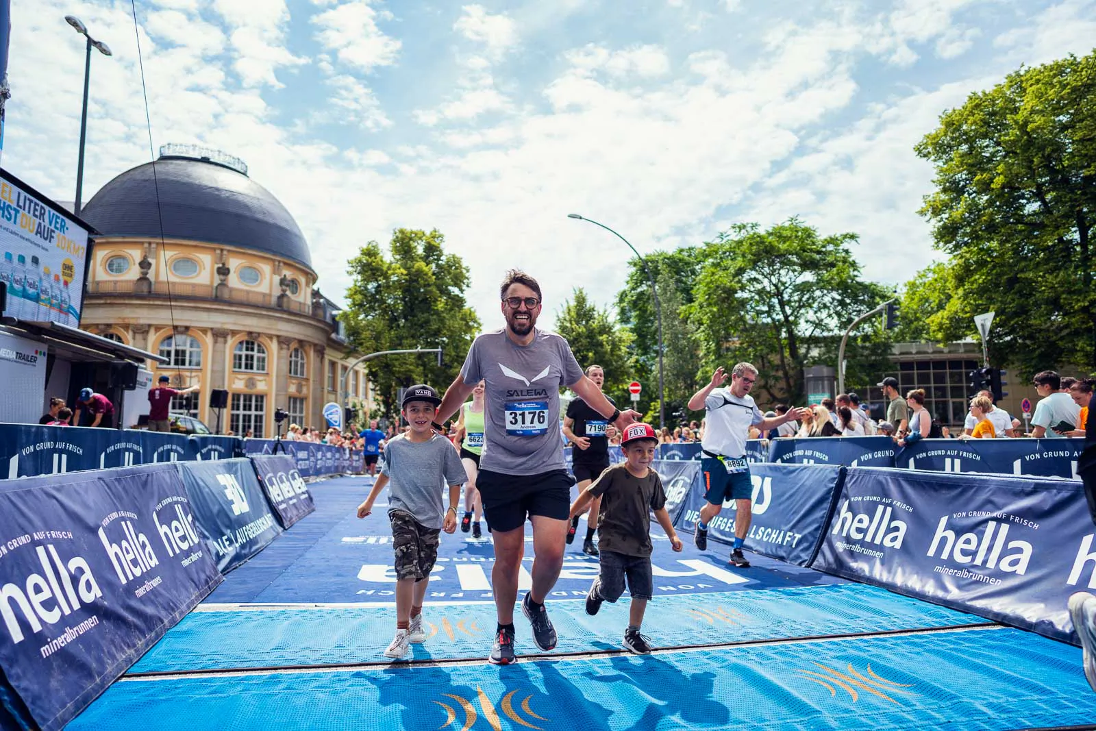 Ein Läufer läuft mit zwei Kindern über die blaue Zielmatte beim Hella Halbmarathon im Sonnenschein.