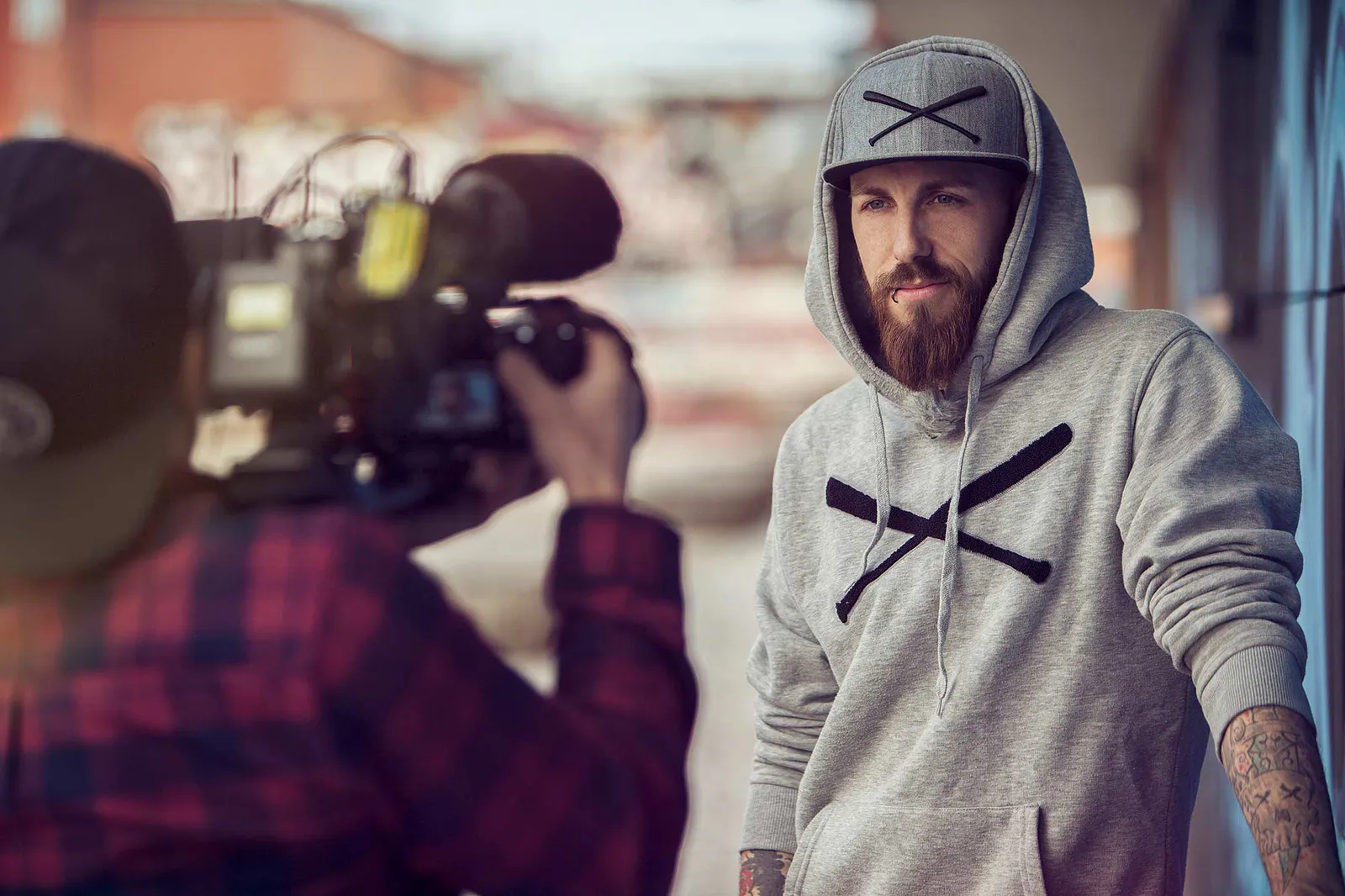 Ein Kameramann filmt Dennis Grundt im grauen Kapuzenpullover und mit Kappe bei Tageslicht vor einer Wand.