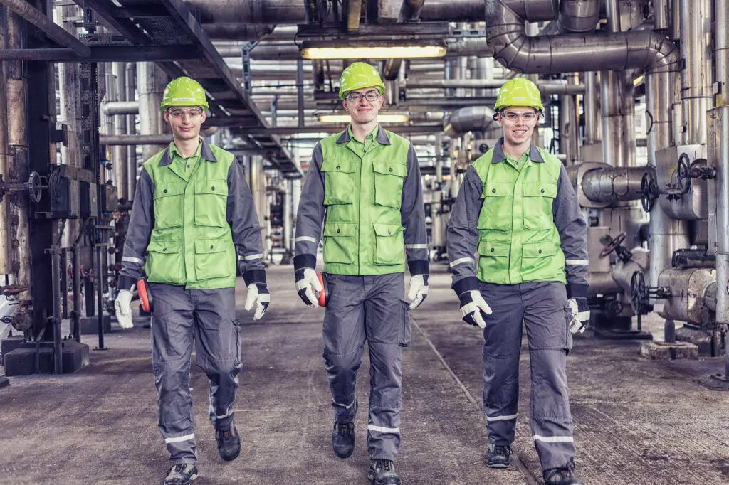 Drei Arbeiter mit grünen Helmen und Westen gehen zwischen Rohrleitungen durch eine Halle der Raffinerie Heide.