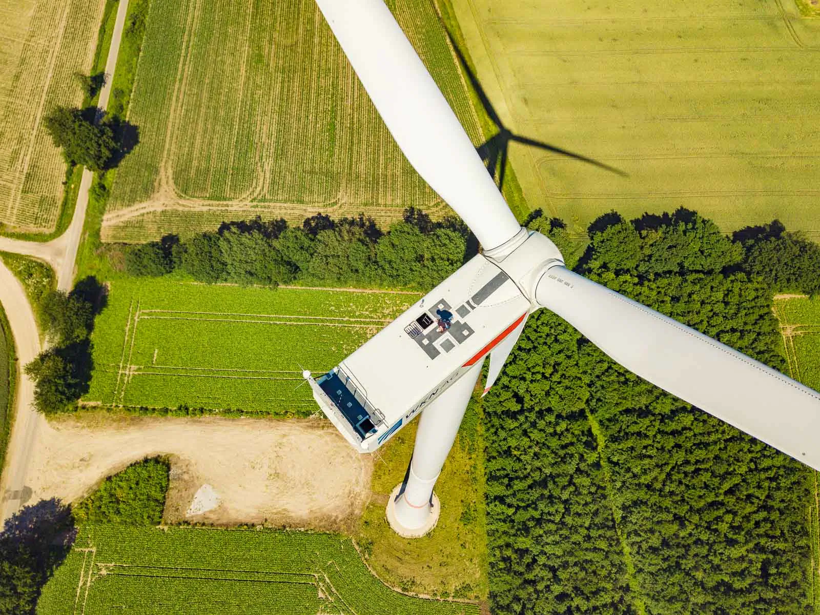 Luftaufnahme einer Windkraftanlage mit weißen Rotorblättern über sonnigen Feldern und einem Waldstück.