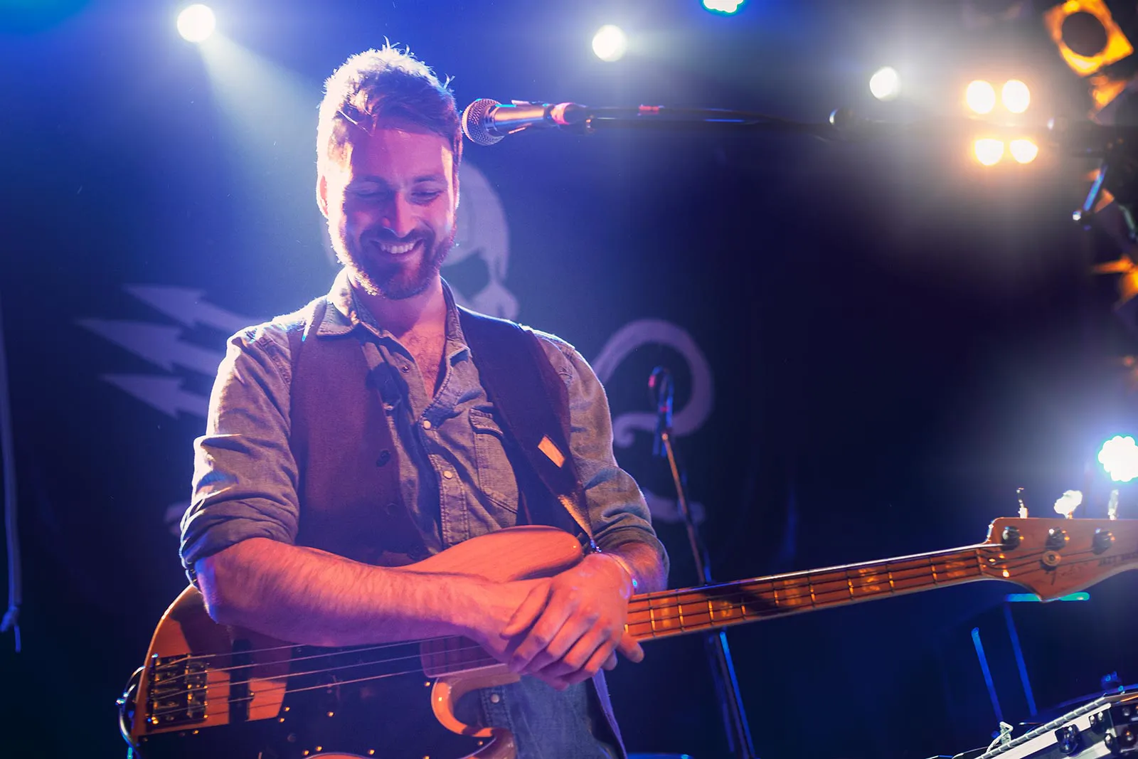 Lächelnder Bassist mit E-Bass vor einem Mikrofon auf einer blau und weiß beleuchteten Konzertbühne.